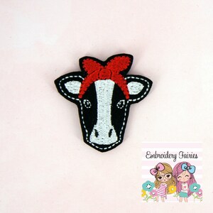 May include: Embroidered patch of a black and white cow with a red bow. The patch is made of felt and has a stitched outline. The text "Embroidery Fairies" is visible in the lower right corner of the image.