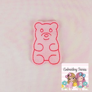 Könnte beinhalten: Ein pinkfarbener, glitzernder Gummibären-förmiger Aufnäher mit roten Nahtdetails. Der Aufnäher hat ein cartoonartiges Design und ist vor einem hellrosa Hintergrund platziert. Das Bild enthält auch das Logo "Embroidery Fairies".