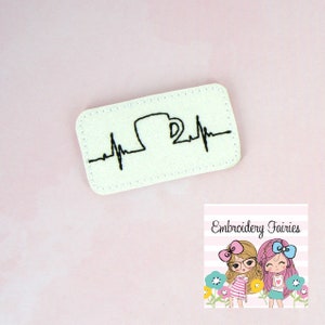 May include: A rectangular, off-white patch with a black embroidered design of a coffee mug within a heartbeat line. The patch is on a pink background. The lower right corner features a logo for Embroidery Fairies.