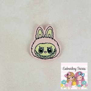 Könnte beinhalten: Rosa gestickter Aufnäher eines Cartoon-Hasen mit einem zahnigen Grinsen und großen Augen. Der Hase trägt einen rosa flauschigen Mantel.