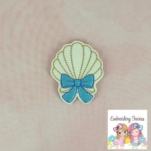 May include: A white seashell-shaped patch with gold glitter accents and a light blue bow. The patch is an embroidered design. The lower right corner of the image has the text "Embroidery Fairies" with a cartoon graphic.