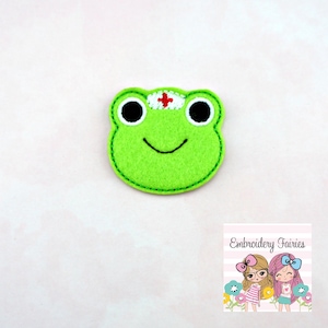 May include: Green felt frog applique with a red cross on its head. The frog is smiling and has black eyes.