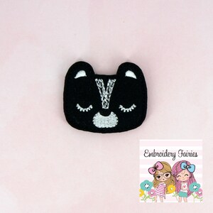 May include: Black felt applique of a cartoon skunk face with white eyes, nose, and stripe. The applique is ideal for adding a whimsical touch to crafts and projects.