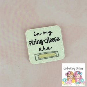May include: A square, off-white embroidered patch with the words "in my string cheese era" in black script. A string cheese graphic is below the text. The patch has a stitched border and is on a pink background.