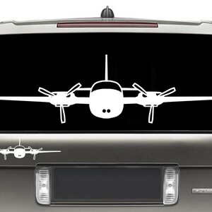 以下が含まれることがあります： 黒い背景に白い小さな飛行機のシルエット。飛行機は翼を広げて前を向いている。飛行機は銀色のSUVのリアウィンドウにある。
