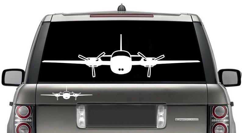 以下が含まれることがあります： 黒い背景に白い小さな飛行機のシルエット。飛行機は翼を広げて前を向いている。飛行機は銀色のSUVのリアウィンドウにある。