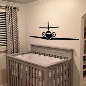 May include: A gray crib with white bedding and a black airplane decal on the wall behind it.