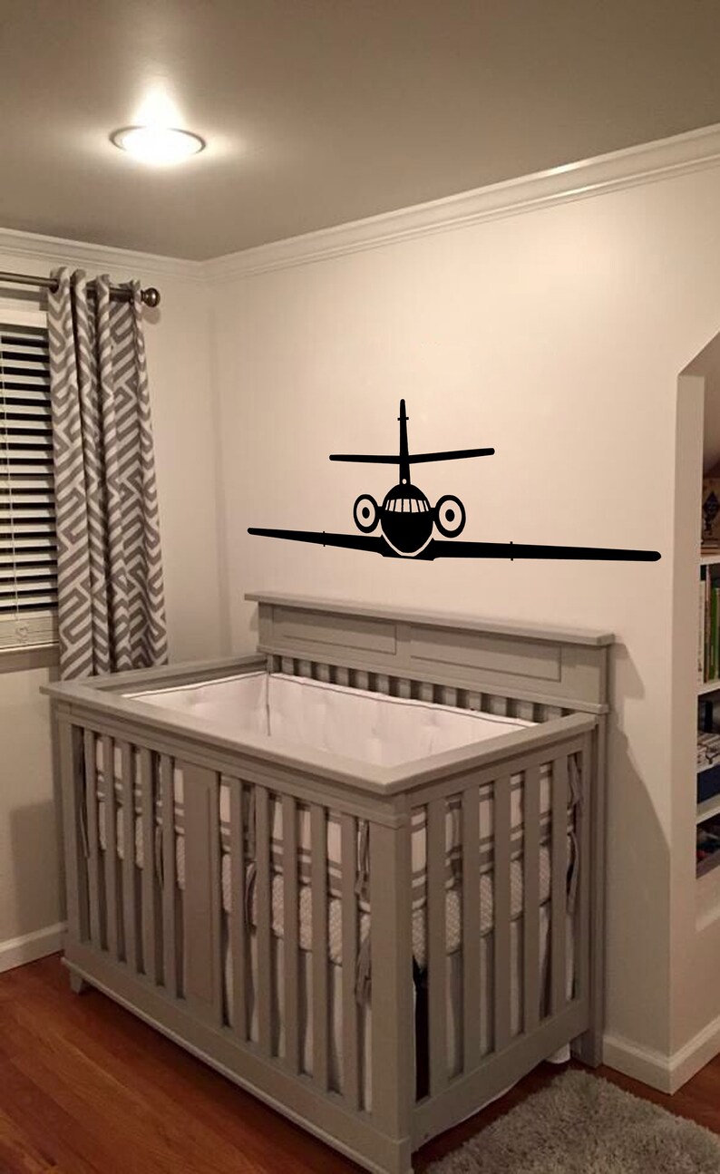 May include: A gray crib with white bedding and a black airplane decal on the wall behind it.