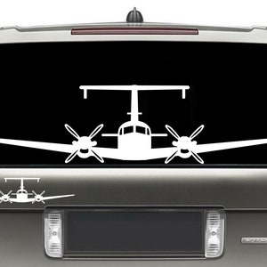 以下が含まれることがあります： 黒い背景に、双発機の白いビニールステッカー。ステッカーは、銀色のSUVのリアウィンドウに貼られています。