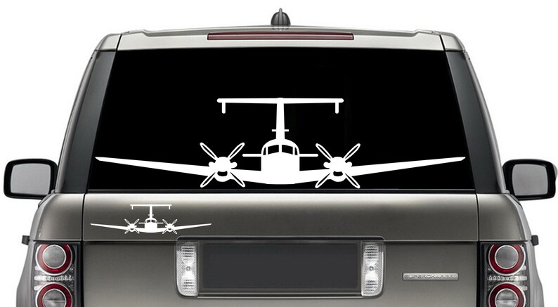 以下が含まれることがあります： 黒い背景に、双発機の白いビニールステッカー。ステッカーは、銀色のSUVのリアウィンドウに貼られています。
