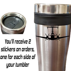 以下が含まれることがあります： 黒い蓋と中央に黒いバンドが付いたステンレス製のトラベルマグ。マグには、前面に小さな飛行機の黒いシルエットが描かれています。「You'll receive 2 stickers on orders, one for each side of your tumbler」という黒いテキストが画像の下に書かれています。
