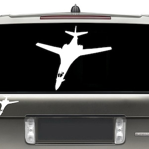 May include: A white silhouette of a B-1B Lancer bomber on a black background. The silhouette is on the back window of a silver SUV.