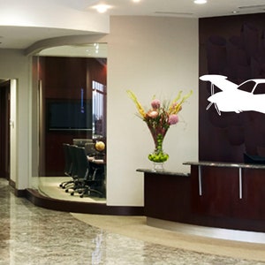 Puede incluir: Una silueta blanca de un avi&oacute;n peque&ntilde;o est&aacute; en una pared marr&oacute;n oscuro en el vest&iacute;bulo de una oficina moderna. El vest&iacute;bulo tiene un mostrador de recepci&oacute;n con un jarr&oacute;n de flores y una puerta de vidrio que conduce a otra habitaci&oacute;n.
