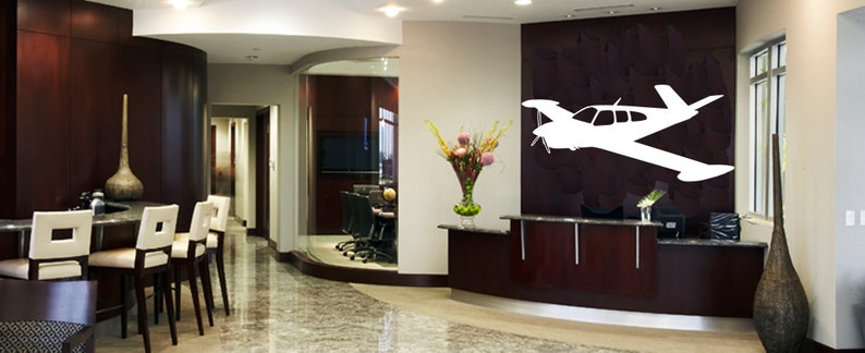 Puede incluir: Una silueta blanca de un avi&oacute;n peque&ntilde;o est&aacute; en una pared marr&oacute;n oscuro en el vest&iacute;bulo de una oficina moderna. El vest&iacute;bulo tiene un mostrador de recepci&oacute;n con un jarr&oacute;n de flores y una puerta de vidrio que conduce a otra habitaci&oacute;n.