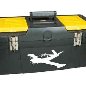 Puede incluir: Una caja de herramientas negra con tapas amarillas y una calcoman&iacute;a de silueta de avi&oacute;n blanco. La caja de herramientas tiene una manija de transporte y dos cierres.