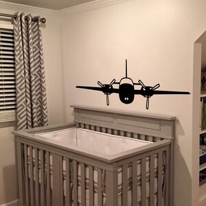 以下が含まれることがあります： 壁に黒い飛行機のステッカーが貼られた、白いシーツを敷いた灰色のベビーベッド