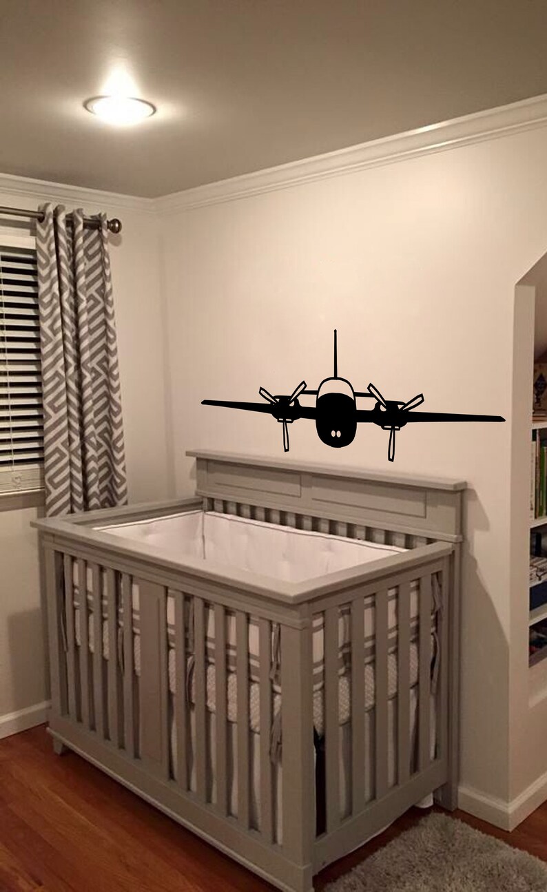 以下が含まれることがあります： 壁に黒い飛行機のステッカーが貼られた、白いシーツを敷いた灰色のベビーベッド