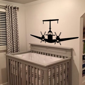 以下が含まれることがあります： 白いシーツを敷いた灰色のベビーベッド。飛行機のシルエットの黒いウォールステッカーが貼られた子供部屋です。