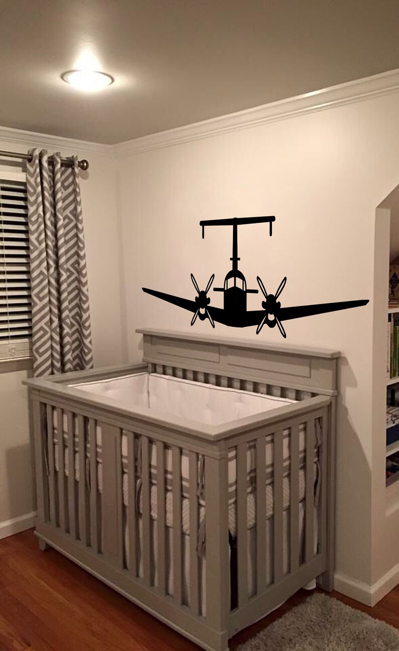 以下が含まれることがあります： 白いシーツを敷いた灰色のベビーベッド。飛行機のシルエットの黒いウォールステッカーが貼られた子供部屋です。