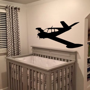 Puede incluir: Una cuna gris con ropa de cama blanca en una habitaci&oacute;n de beb&eacute; con una silueta negra de un avi&oacute;n peque&ntilde;o en la pared.