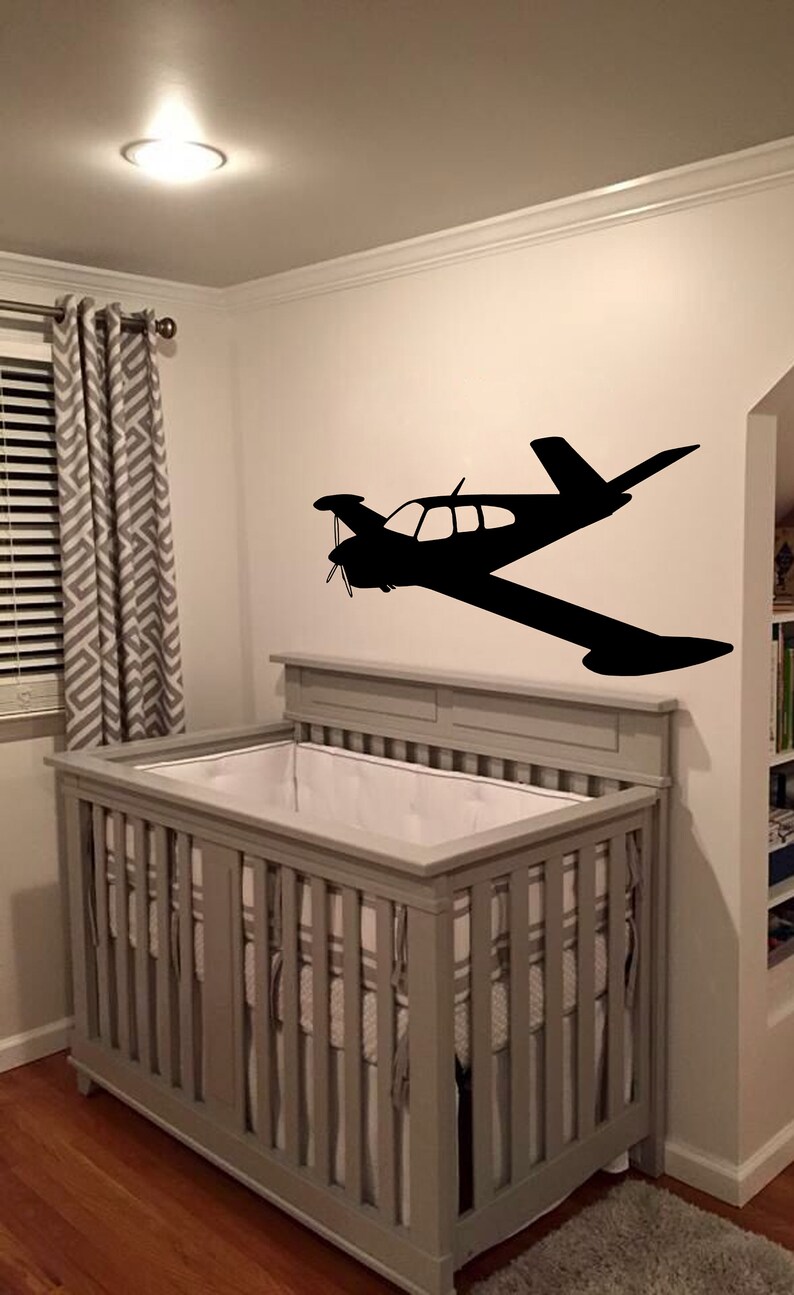 Puede incluir: Una cuna gris con ropa de cama blanca en una habitaci&oacute;n de beb&eacute; con una silueta negra de un avi&oacute;n peque&ntilde;o en la pared.