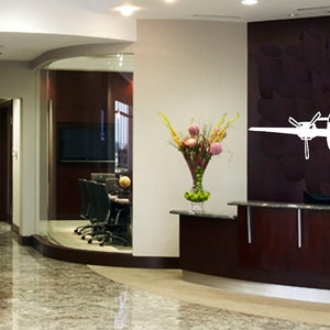 以下が含まれることがあります： 受付カウンターと壁に大きな白い飛行機のステッカーが貼られた、モダンなオフィスのインテリア。オフィスはダークウッドのパネリングと、受付エリアと会議室を隔てるガラスの壁がある。