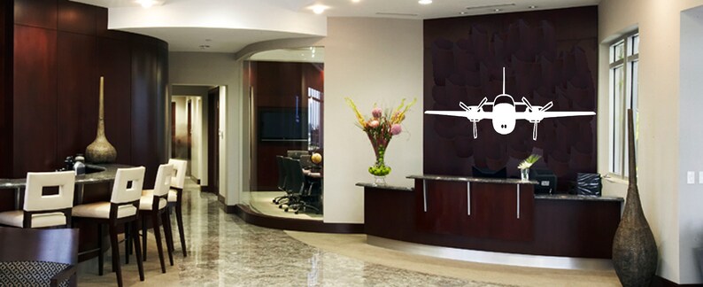 以下が含まれることがあります： 受付カウンターと壁に大きな白い飛行機のステッカーが貼られた、モダンなオフィスのインテリア。オフィスはダークウッドのパネリングと、受付エリアと会議室を隔てるガラスの壁がある。