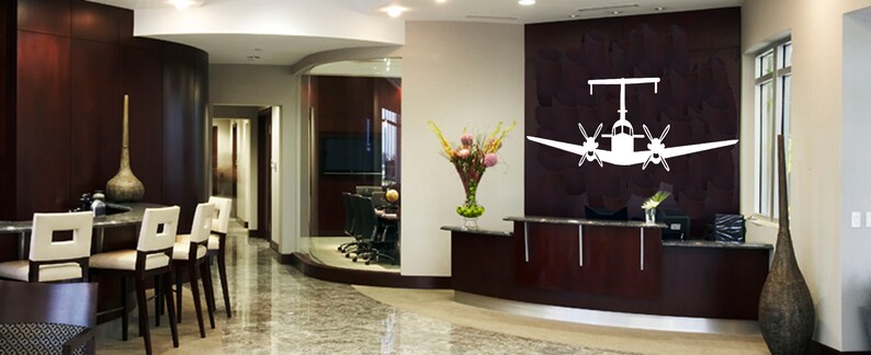 以下が含まれることがあります： 受付カウンターと壁に白いシルエットの小型飛行機が描かれた、モダンなオフィスロビー。オフィスはダークウッドのパネリングと、ロビーと会議室を隔てるガラスの壁がある。