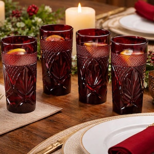 Puede incluir: Cuatro vasos de beber de color rojo rubí, estampados, llenos de una bebida y una rodaja de cítricos, colocados sobre una mesa de madera. Los vasos forman parte de una mesa con velas, platos y cubiertos dorados. Una escena de comedor festiva y elegante.
