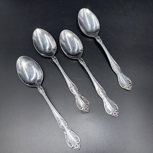 May include: Four silver spoons with ornate handles are arranged diagonally on a dark surface. The spoons have a classic design with a rounded bowl and detailed floral patterns on the handles, suggesting a vintage or antique style.