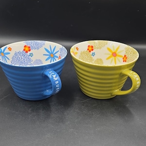 Puede incluir: Dos tazas de cerámica acanaladas con la palabra Starbucks grabada en las asas. La taza azul presenta un patrón interior de flores azules con centros naranjas. La taza verde lima tiene un patrón interior de flores amarillas con centros naranjas.