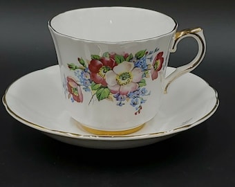 May include: White teacup and saucer set with a floral design featuring pink, red, and blue flowers. The cup and saucer have gold trim. The teacup is sitting on the saucer.