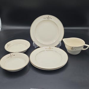 May include: A set of cream-colored ceramic dinnerware featuring a gold-toned Masonic square and compass emblem with the text Atchison Kan. The collection includes two plates, two shallow bowls, and a creamer pitcher, all with thin gold trim.