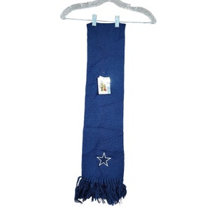 May include: A blue knitted scarf with a white star and a fringe at the bottom. The scarf has a tag that says "Dallas Cowboys".