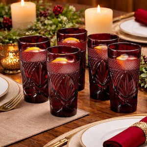 May include: Six ruby red drinking glasses filled with a beverage and a slice of citrus. The glasses have a textured leaf and geometric design. Lit candles and decorative greenery are in the background, suggesting a festive occasion.