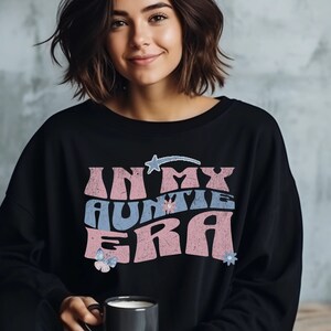 May include: A black sweatshirt with a pink and blue graphic that says "In My Auntie Era" with a star and flowers.