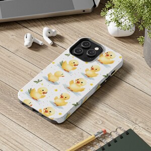 May include: A white iPhone case featuring a pattern of yellow ducklings on a white background. The case has a camera lens cutout and a white background.