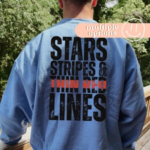 Może przedstawiać: Błękitna bluza z napisem "STARS, STRIPES & THIN RED LINES" w postarzanym czarnym kroju pisma, z "THIN RED" w kolorze czerwonym. Słowa "multiple options" znajdują się nad uśmiechniętą buzią.