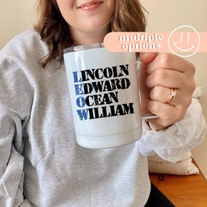 Peut inclure: Mug de voyage blanc avec les noms "LINCOLN", "EDWARD", "OCEAN" et "WILLIAM" imprimés en bleu et noir. Le mug a un bord argenté et une anse blanche. Une bannière rose indique "multiple options".