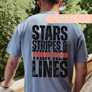 Może przedstawiać: Jasnoniebieska koszulka z napisem "STARS, STRIPES & THIN RED LINES" w kolorze czarnym i czerwonym. Koszulka ma postarzany, vintage wygląd. Tekst jest ułożony pionowo.