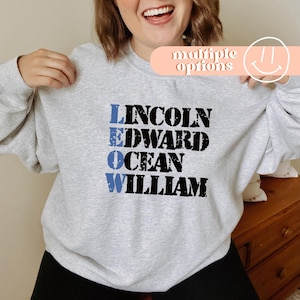 May include: A light gray sweatshirt with the names "LINCOLN", "EDWARD", "OCEAN", and "WILLIAM" printed in a distressed font. The names "LINCOLN" and "OCEAN" are in blue, while "EDWARD" and "WILLIAM" are in black. The words "multiple options" are above the sweatshirt.