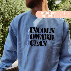 May include: A light blue sweatshirt with the words "LINCOLN EDWARD OCEAN" printed in a layered design. The text is in a mix of blue and black. The top right corner has a peach-colored banner with the words "multiple options" and a smiley face.