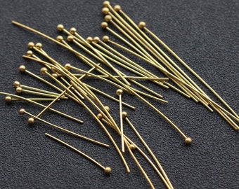 Op de afbeelding: Een stapel gouden kopspelden met ronde koppen. De spelden liggen verspreid op een donkergrijs oppervlak.