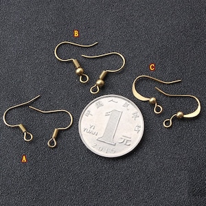 May include: Three gold-toned earring hooks, each with a small ball at the end. The hooks are arranged in a row on a dark surface, with a Chinese coin in the centre.