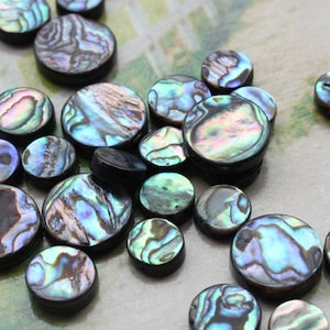 Pode incluir: Uma coleção de botões redondos de concha de abalone iridescente com suporte preto. Os botões são de vários tons de azul, verde e roxo.