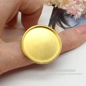 Puede incluir: Un anillo dorado con una superficie grande, redonda y plana. El anillo se sostiene en un dedo, mostrando su diseño. El anillo tiene un estilo simple y elegante, adecuado para varios proyectos de artesanía o como un accesorio único.