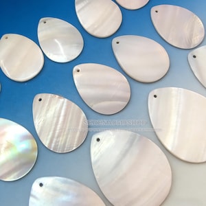 May include: A collection of pearlescent, teardrop-shaped pendants with a small hole at the top for stringing. The pendants have a natural, iridescent sheen and are arranged on a gradient blue background. The pendants are likely for jewelry making.