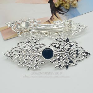 May include: Two silver hair barrettes with detailed filigree patterns. One has a hinged clasp, the other a dark blue circular centre. The barrettes are decorative hair accessories, suitable for adding a touch of elegance to hairstyles.