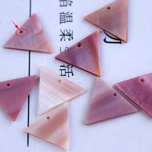 Puede incluir: Siete colgantes de concha de forma triangular de color rosa y blanco con un agujero en el centro de cada uno. Los colgantes están sobre una superficie blanca.