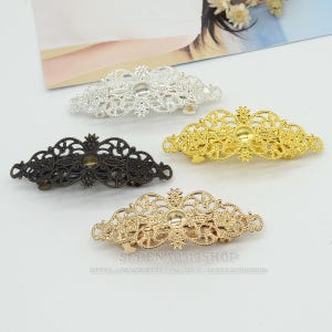 Könnte beinhalten: Vier verzierte Haarspangen aus Metall in Silber, Gold, Bronze und Antikgold. Jede Spange hat ein filigranes Design mit einem zentralen kreisförmigen Schmuckelement. Die Spangen werden auf einer weißen Oberfläche präsentiert.